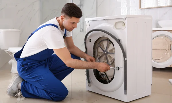 Réparateur de machine à laver à Paris, vêtu d'un bleu de travail, agenouillé derrière une machine à laver blanche dans une salle de bain