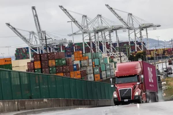 Un camion rouge tracteur remorque transportant un conteneur maritime circule le long d'une route adjacent au terminal portuaire d'Oakland, en Californie, avec de grandes grues et des conteneurs colorés en arrière-plan