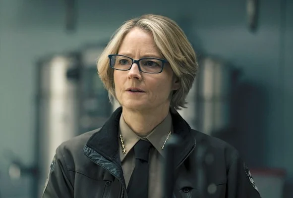 Jodie Foster, cheveux courts blonds et lunettes, portant une veste sombre avec chemise et cravate, arborant un regard sérieux dans True Detective Night Country