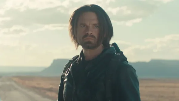 Sebastian Stan, acteur de Bucky Barnes, portant une veste sombre avec cheveux longs et barbe, debout dans un paysage désertique bordé d'une route et de mesas en arrière-plan