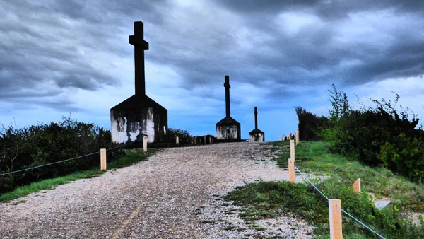 Chemin de gravier montant vers trois grandes croix sombres en silhouette sous un ciel nuageux, à la montagne des Trois Croix de Santenay, bordé de poteaux et de cordage en bois