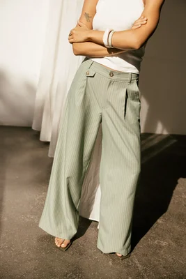 Pantalon Alison Looney à pinces rayé vert clair et large, porté avec un haut blanc sans manches, porté par une femme les bras croisés sur fond neutre