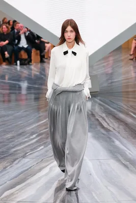 Mannequin féminin défilant sur un podium en pantalon gris à pinces et taille haute, portant un chemisier blanc à nœud noir