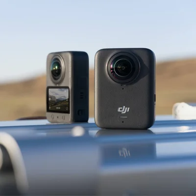 Deux caméras d'action DJI Osmo 360 posées sur une surface bleue en extérieur : l'une noire avec lentille et logo DJI en premier plan, l'autre grise avec un écran affichant un paysage en arrière-plan