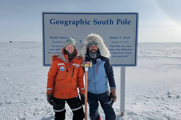 Deux explorateurs en combinaison polaire lourde posant devant la marque du Pôle Sud géographique en Antarctique après leur traversée en kite-ski