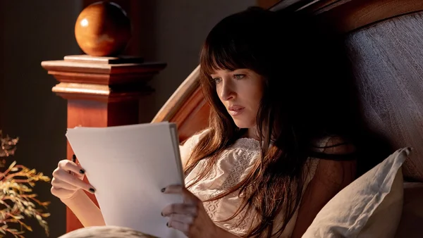 Dakota Johnson, longue chevelure brune et frange, assise dans un lit et lisant un livre, éclairée par une lumière chaude et tamisée.