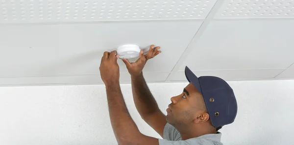 Un homme portant une casquette de baseball bleue et un t-shirt gris, bras levé, installe ou ajuste un détecteur de fumée blanc et circulaire sur un plafond suspendu