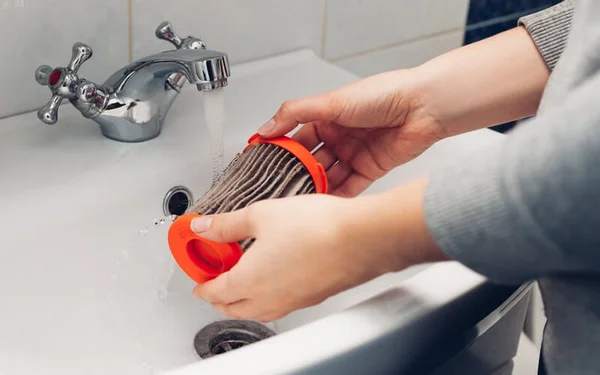 Mains rincant un filtre plissé cylindrique à extrémités orange sous un robinet dans un lavabo blanc
