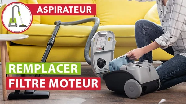 Personne à genoux sur un parquet en bois remplaçant le filtre moteur d'un aspirateur à sac gris à côté d'un canapé jaune