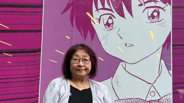 Rumiko Takahashi, mangaka japonaise aux lunettes et cheveux sombres, se tenant devant une grande fresque d'art anime représentant le visage d'un personnage