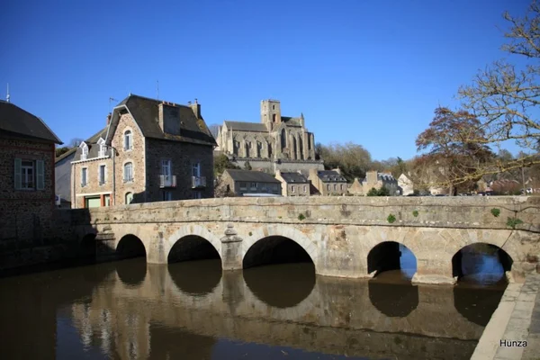 Vue de Lamballe depuis la rivière : le pont de pierre à plusieurs arches, les maisons anciennes et, sur la colline, la silhouette gothique de la collégiale sous un ciel bleu dégagé.