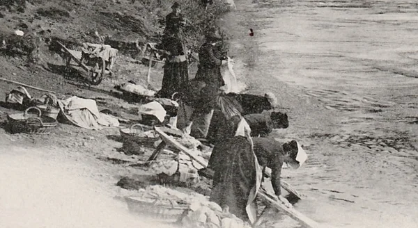 Photographie ancienne en noir et blanc montrant un groupe de lavandières agenouillées au bord d'une rivière, battant le linge à la main sur des planches de bois, avec des paniers posés sur la berge.