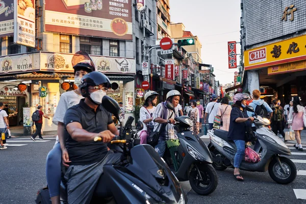 Scène de rue animée à Taipei avec plusieurs personnes à scooter arrêtées à un carrefour, des piétons et des devantures de commerces en arrière-plan