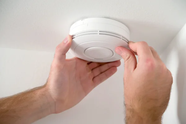 Vue rapprochée en contre-plongée des mains d'une personne agrippant les côtés d'un détecteur de fumée blanc et circulaire pour l'installer ou le vérifier sur un plafond blanc