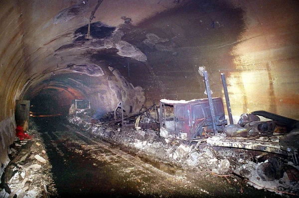 Intérieur du tunnel du Mont-Blanc après l'incendie de 1999 : parois et plafond noircis par la suie, carcasses de véhicules calcinées et débris éparpillés le long de la paroi, éclairé depuis la droite.