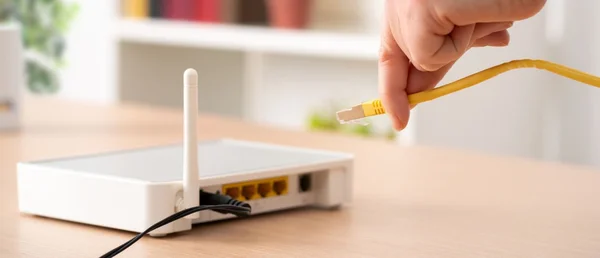 Une main tenant un câble Ethernet jaune sur le point de le brancher dans un routeur Wi-Fi blanc posé sur une table en bois