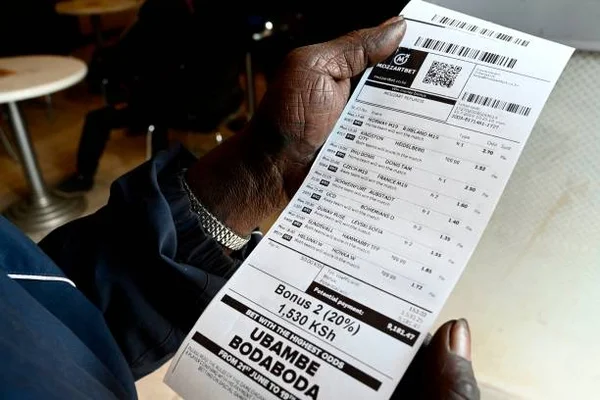Main tenant un ticket de paris sportifs Mozzartbet affichant des matchs, des cotes et un gain potentiel de 1 530 KSh dans un bar de paris à Nairobi, au Kenya
