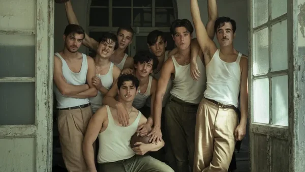 Des jeunes hommes vêtus de maillots de corps blancs et de pantalons clairs posent ensemble dans l'embrasure d'une porte, l'un d'eux portant un ballon de football