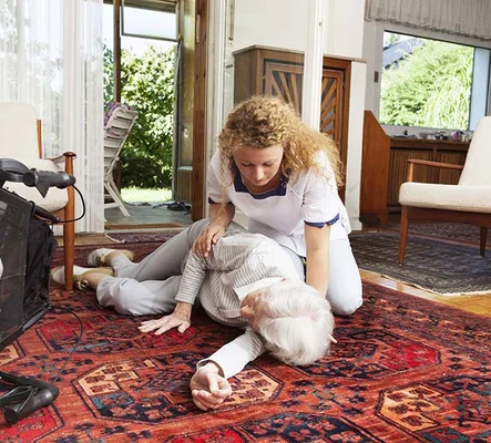 Une infirmière libérale en tenue blanche, agenouillée sur un tapis à motifs, porte assistance à une personne âgée allongée au sol dans son domicile pendant une urgence.