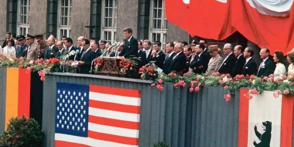 John Fitzgerald Kennedy prenant la parole depuis une tribune officielle ornée des drapeaux de l'Allemagne, des États-Unis et de Berlin, lors de son discours « Ich bin ein Berliner » devant le mur de Berlin.