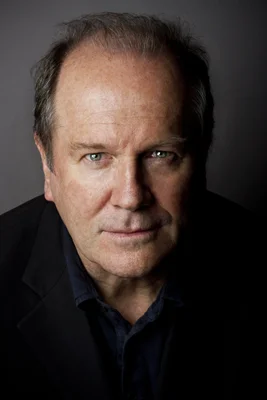 Portrait studio de l'écrivain britannique William Boyd, cheveux clairsemés et veste sombre sur chemise foncée, regard tourné vers l'objectif sur fond noir
