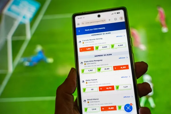 Main tenant un smartphone affichant les cotes d'un match de Coupe du monde sur une application de paris sportifs, sur fond flou de terrain de football