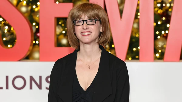 Freida McFadden, cheveux courts brun et lunettes, en blazer noir, debout devant un décor rose portant de grandes lettres lors d'un événement