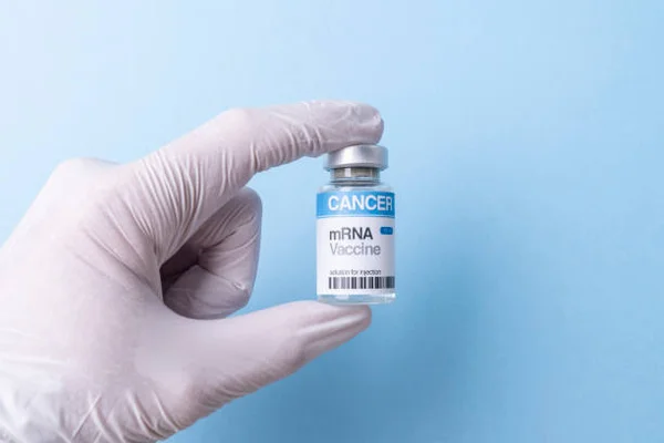 Main gantée de blanc tenant un petit flacon de verre étiqueté « CANCER mRNA Vaccine » devant un fond bleu clair uni