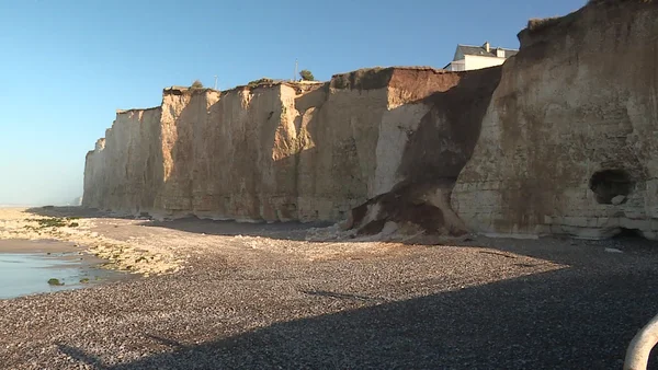 Falaise crayeuse en cours d'érosion en Normandie, surplombant une plage de galets sous un ciel bleu dégagé, avec une habitation construite au bord du sommet menacé d'effondrement