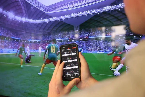 Une personne tient un smartphone ouvert sur une application de paris sportifs, un match de football diffusé sur grand écran en arrière-plan