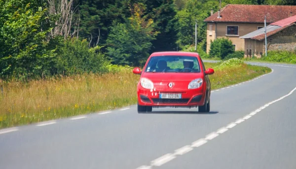 Une Renault Clio RS rouge roule sur une route de campagne sinueuse bordée d'arbres, avec une maison en arrière-plan.