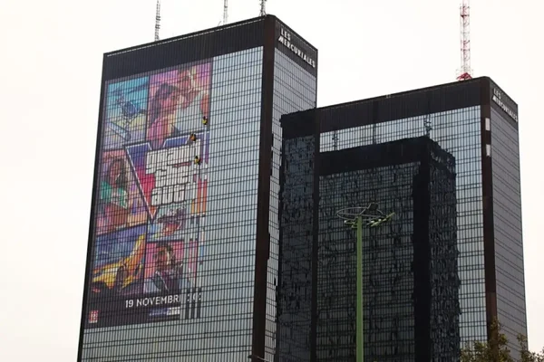Les tours Mercuriales, deux gratte-ciel de verre sombre, avec une immense bannière publicitaire colorée pour GTA 6 recouvrant la façade de la tour de gauche sous un ciel couvert
