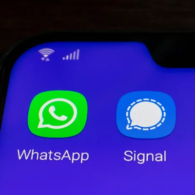 Gros plan sur l'écran d'un smartphone où apparaissent côte à côte les icônes des applications de messagerie WhatsApp et Signal, sur un fond bleu uni