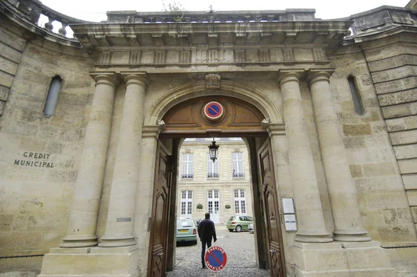 Le grand portail en pierre du Crédit municipal de Bordeaux, ouvert sur une cour intérieure où stationnent des voitures, une personne franchissant l'entrée sous un panneau d'interdiction de stationner.