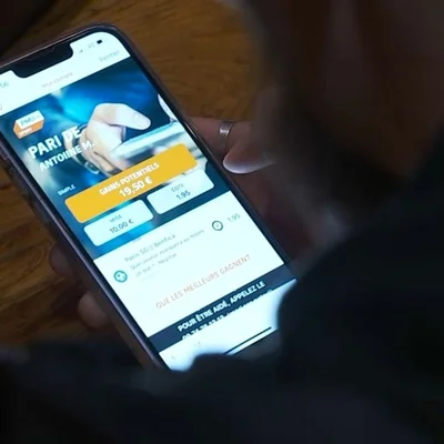 Gros plan sur un smartphone tenu à la main affichant une application de paris sportifs en ligne avec des cotes et des gains potentiels, illustration de l'addiction des jeunes joueurs