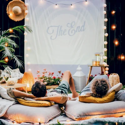 Un couple allongé sur des coussins dans un jardin regarde un film projeté sur un drap blanc affichant « The End », entouré de guirlandes lumineuses et de plantes