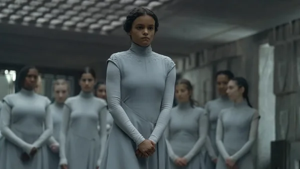 Une jeune femme aux cheveux noirs, vêtue d'une robe bleue claire à col montant, se tient au premier plan dans une salle industrielle sombre avec un groupe de femmes similairement habillées derrière elle.