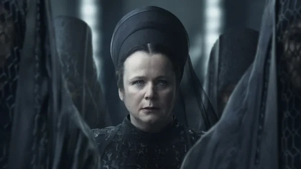 Une femme au visage sévère, vêtant un headpiece sombre structuré et une robe noire texturée, se tient entre deux figures voilées dans une scène sombre inspirée de Dune : Prophecy.