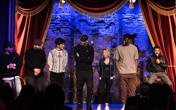 Sept humoristes debout côte à côte sur la scène d'un comedy club parisien, devant un rideau rouge et un mur de pierre éclairé en bleu, lors d'une représentation de stand-up à Paris