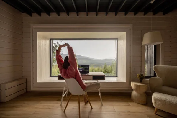 Teletravailur en hoodie rose s'étirant dans une pièce en bois face à une fenêtre donnant sur des montagnes et des arbres, illustrant le télétravail à la campagne
