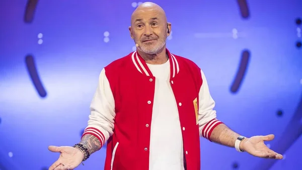 Vincent Lagaf', crâne rasé et barbe grisonnante, vêtu d'une veste universitaire rouge et blanche sur un t-shirt blanc, bras ouverts et paumes vers le ciel sur la scène du « Bigdil », micro-casque sur la tête et tatouages visibles sur l'avant-bras droit.