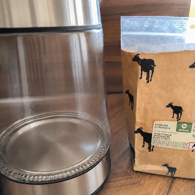 Photo d'une bouilloire électrique en acier inoxydable et verre posée à côté d'un sachet de kyselina citronova (acide citrique) avec des silhouettes de chèvres
