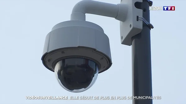 Gros plan sur une caméra de vidéosurveillance en dôme grise fixée à un poteau, sur fond de ciel dégagé, dans un reportage télévisé.