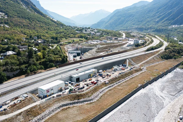 Vue aérienne d'un vaste chantier dans une vallée alpine, avec routes à plusieurs voies, zones de stockage et lit de rivière, pour l'entrée du tunnel de base du Lyon-Turin.