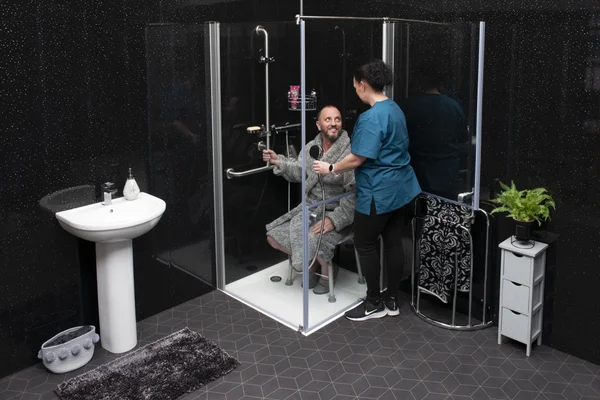Un homme en peignoir assis sur un siège de douche dans une cabine en verre, assisté par une auxiliaire de vie en tenue médicale bleu-vert, dans une salle de bains aux murs sombres au sol en mosaïque hexagonale.