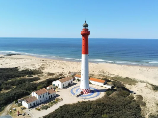 Vue aérienne du phare de la Coubre à La Tremblade, photographe depuis le détroit de Gascogne, montrant le phare rayé rouge et blanc sur une côte sablonneuse entouré de bâtiments et de végétation, menacé par l'érosion marine.