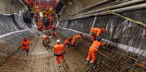 Ouvriers en gilets orange et casques travaillant sur un important ferraillage à l'intérieur d'un tunnel, sur le chantier CO8 du Lyon-Turin, avec engins en arrière-plan.