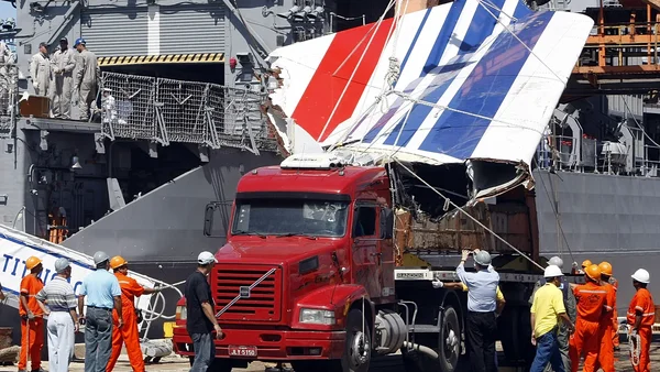 Section endommagée de fuselage d'avion avec les couleurs de l'Air France (rouge, blanc, bleu) étant chargée sur une camionnette rouge par des ouvriers en combinaison de sécurité, avec un grand navire gris en fond.