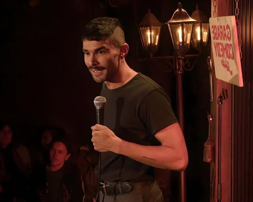 Un humoriste moustachu, cheveux courts et boucle d'oreille, micro en main, se produit en stand-up sur la scène du Garage Comedy Club à Marseille devant un public dans la pénombre