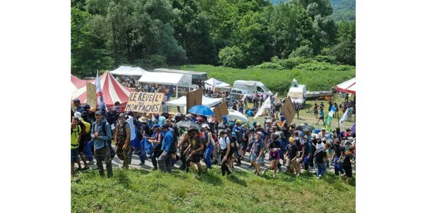 Une foule de manifestants contre le Lyon-Turin marche sur un chemin herbeux en lisière de bois, avec pancartes, drapeaux, tentes et véhicules en arrière-plan.
